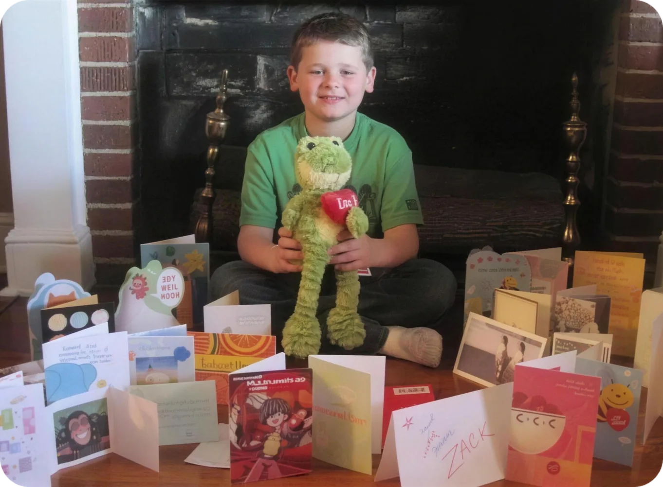 A boy holding a stuffed frog, sitting in front of a fireplace with the cards he received